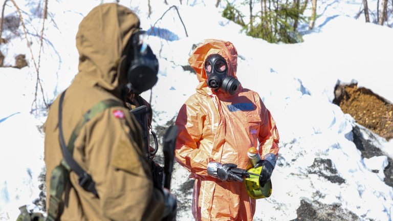 To mennesker med gassmaske og vernedrakt står ute i snødekt landskap med teknisk utstyr.