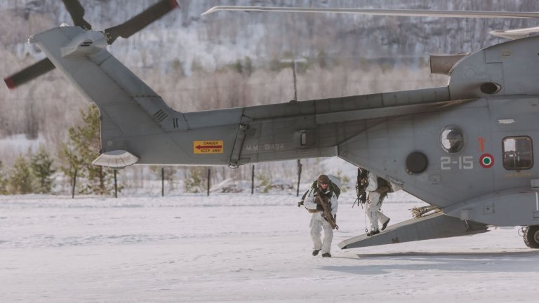 Bevæpnede soldater ikledd vinterkamuflasje på vei ut av et helikopter i snøkledd landskap.