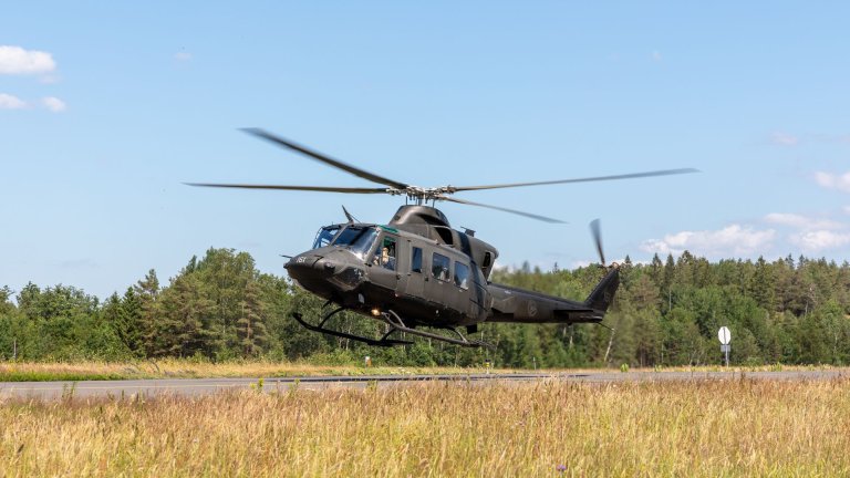 Militærhelikopter under landing på en rullebane med skog og blå himmel i bakgrunnen.