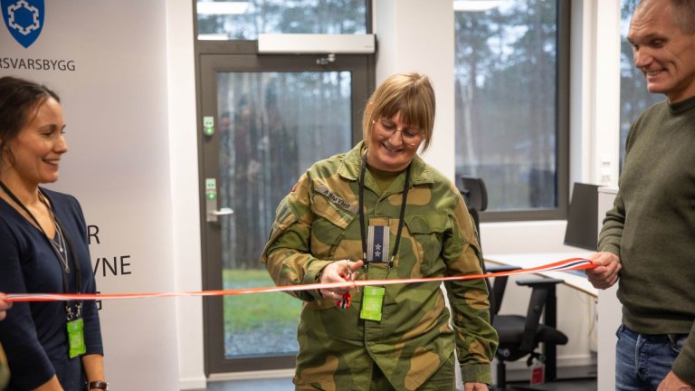 Kvinne i uniform klipper smilende over en snor i rødt, hvitt og blått, som blir holdt av to menn. Kontorlandskap og vinduer i bakgrunnen.