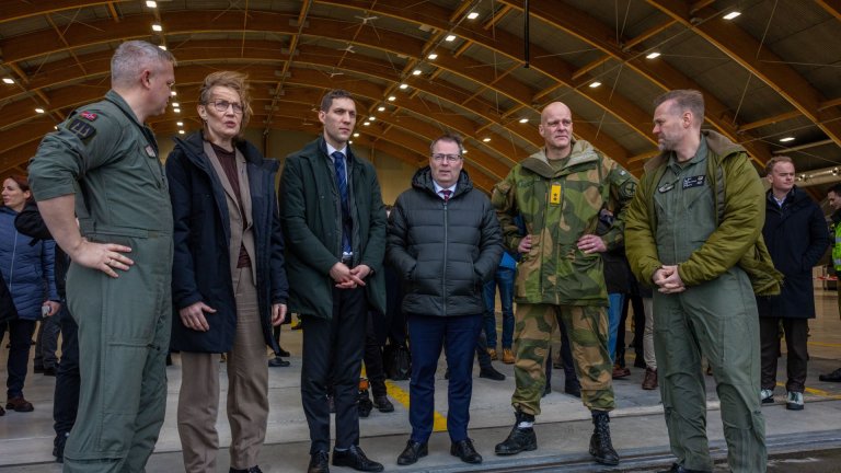 En gruppe personer, inkludert militært personell i uniform og sivile i formelle klær, står sammen i en stor hangar med en buet tak.