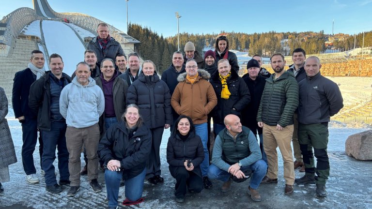 En gruppe mennesker kledd i vinterklær står samlet utendørs foran en hoppbakke. Det er snø og is på bakken, og bakgrunnen viser skogkledde åser og blå himmel.